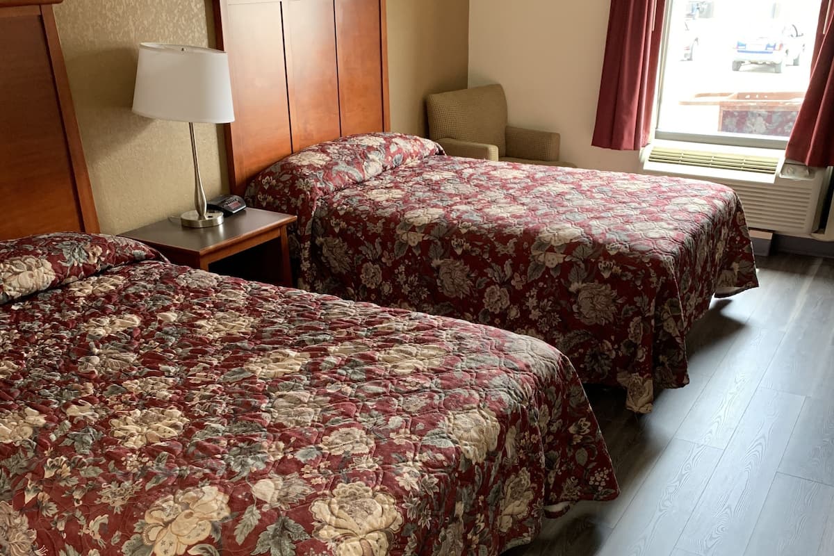 A hotel room with two beds, a wooden headboard, a nightstand with a lamp, a window with curtains, and a radiator.