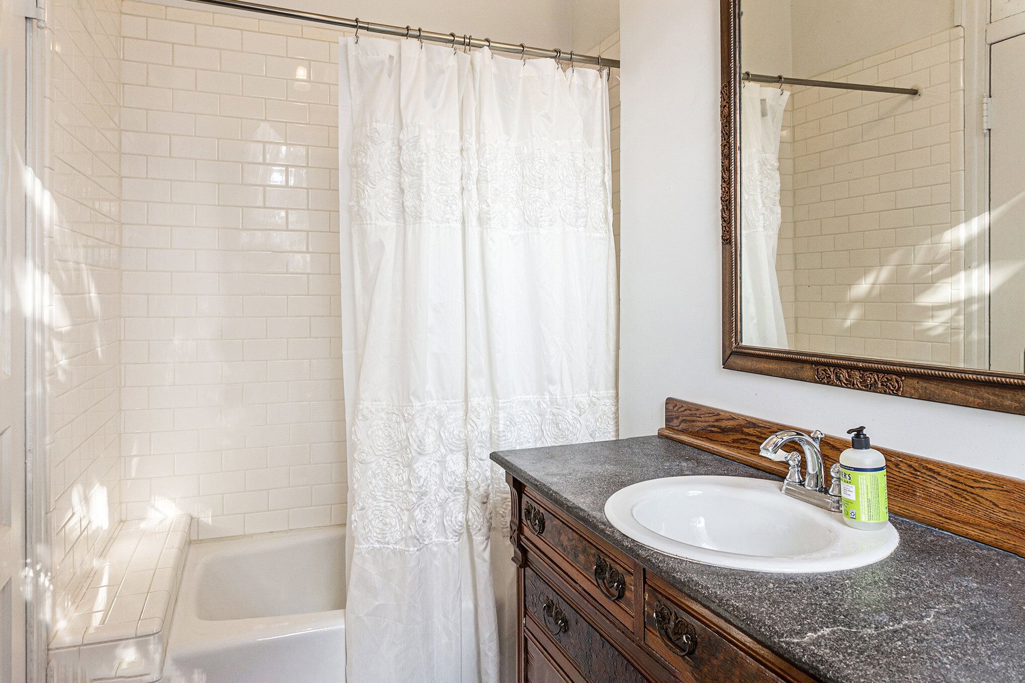 Un baño con una bañera blanca, una cortina de ducha, un lavabo con dispensador de jabón y un espejo con marco de madera.