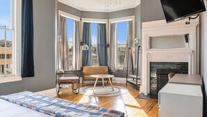 A hotel room with a bed, a seating area with a table, a fireplace, and large windows with curtains.