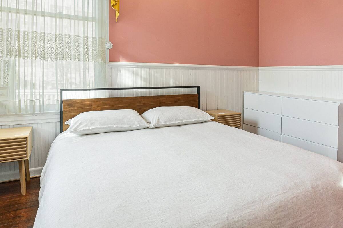 Un lit bien fait, avec une tête de lit en bois, dans une pièce aux murs roses et à la commode blanche.