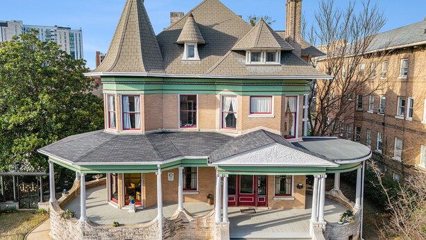 A Victorian-style house with a wrap-around porch, a brick facade, and a steeply pitched roof.