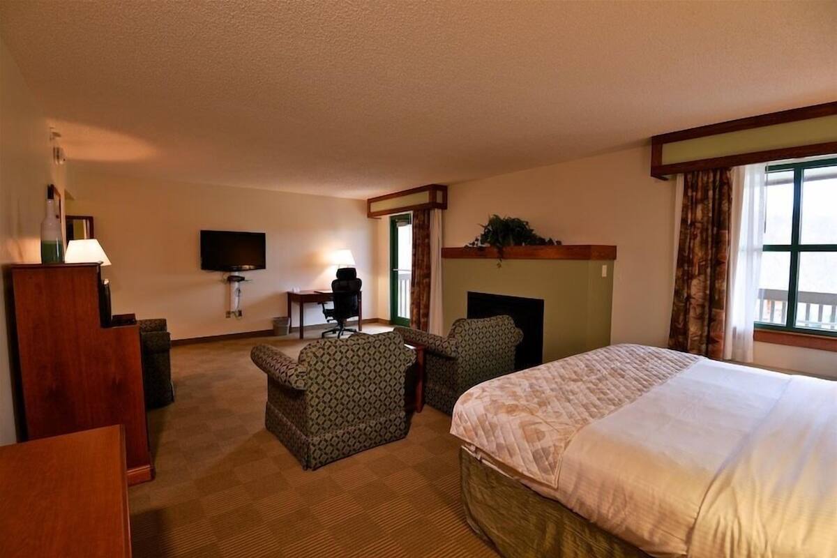 A hotel room with a large bed, a desk, a chair, a television, and a fireplace.
