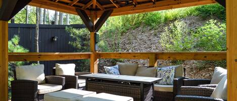 Patio esterno con mobili in vimini e un tavolo, circondato da un muretto in legno e un'area coperta con tetto in legno.