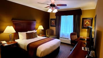 Una habitación de hotel con una cama, dos lámparas de noche, una silla, un escritorio con una computadora, un ventilador de techo y una ventana con cortinas.