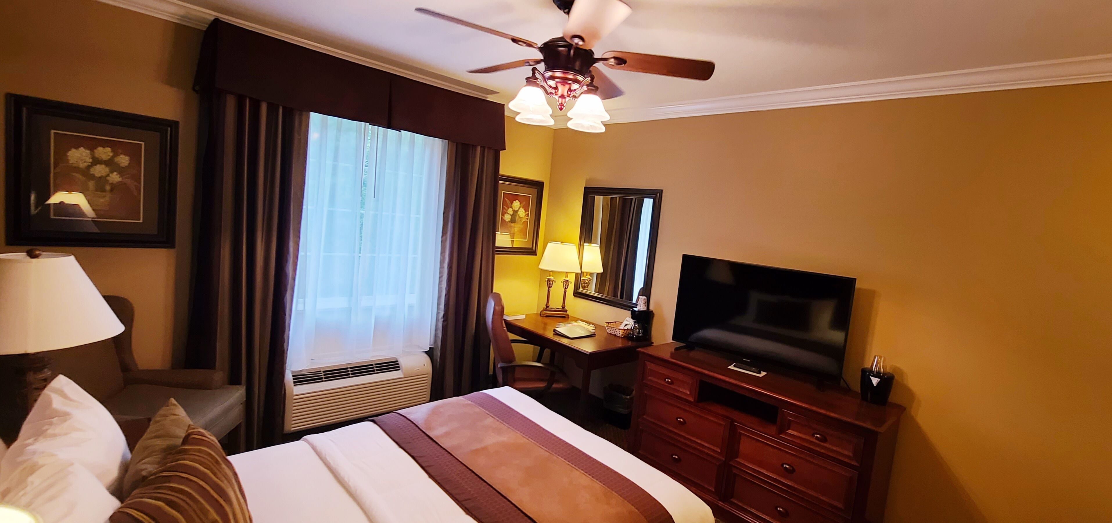 Une chambre d’hôtel comprenant un lit, un bureau avec une chaise, une télévision, un ventilateur de plafond et une fenêtre avec des rideaux.