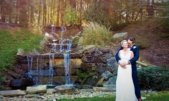 Una pareja parada frente a una cascada, con un muro de piedra y follaje al fondo.