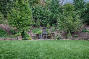 Un jardín ajardinado con una pequeña cascada, rodeado de árboles densos y un muro de contención de piedra.