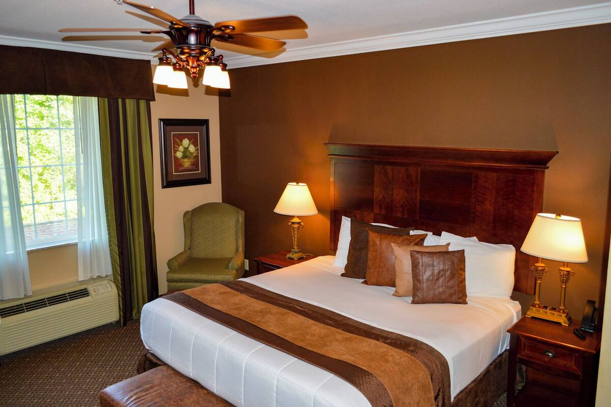 A hotel room with a large bed, two bedside tables with lamps, a ceiling fan, a window with curtains, and a framed picture on the wall.