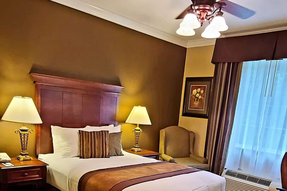 A hotel room with a large bed, two bedside lamps, a ceiling fan, a chair, and a window with curtains.