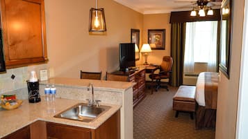 A hotel room with a kitchenette, a desk with a chair, a bed, and a ceiling fan.