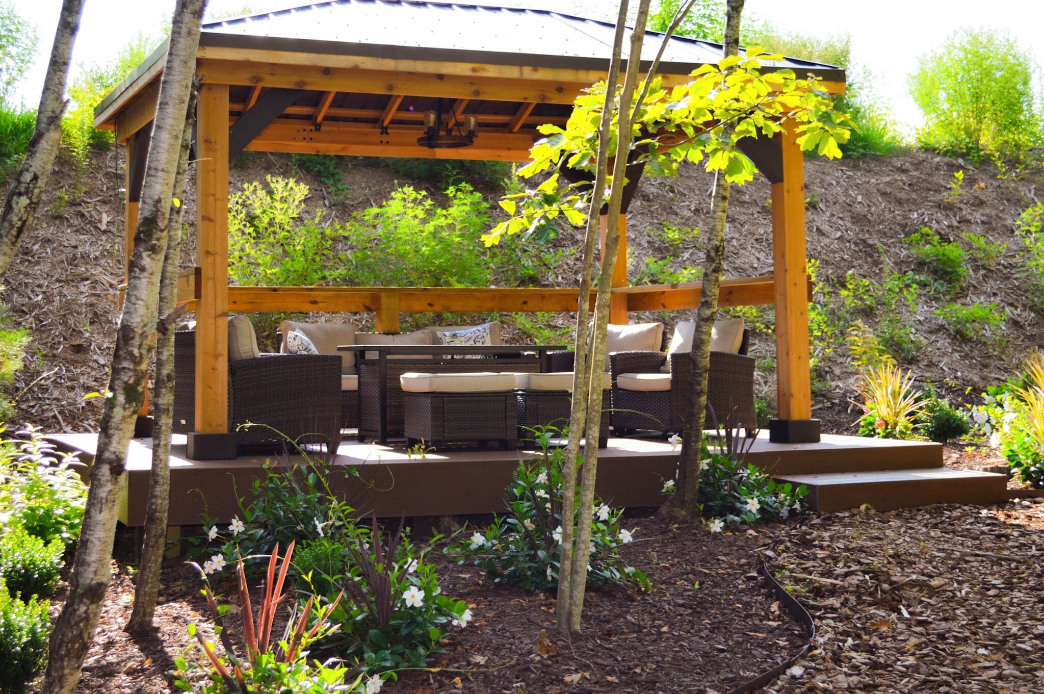 A wooden pergola with a built-in seating area, surrounded by a landscaped garden with various plants and flowers.