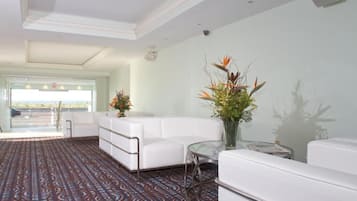 A hotel lobby with a patterned carpet, white sofas, a glass table, and a large window with a view.
