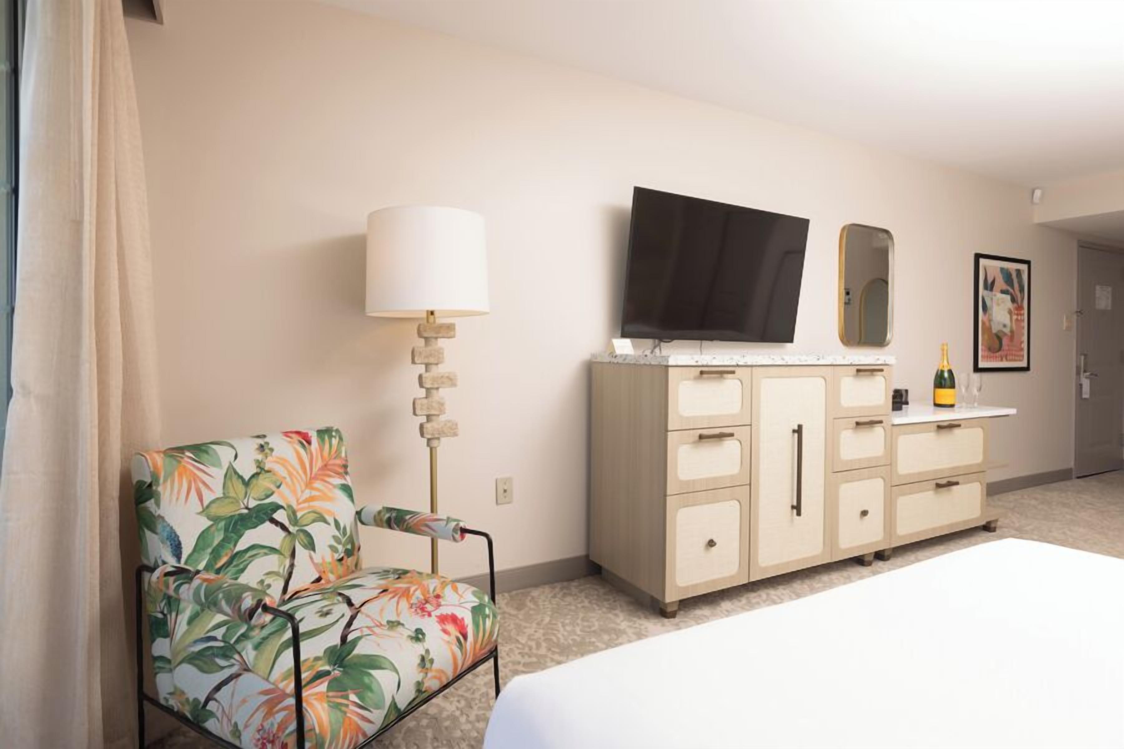 Una habitación de hotel con una cama, un sillón floral, un aparador, un televisor y un espejo.