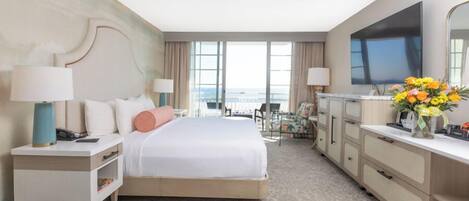 A hotel room with a large bed, bedside tables, a desk, a chair, a flat-screen TV, and a view of the ocean.