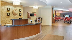 Un'area reception di un hotel con una scrivania a reception in legno curvata, una parete in pietra con un motivo a mezzaluna e una zona salotto con sedie e tavoli rossi.
