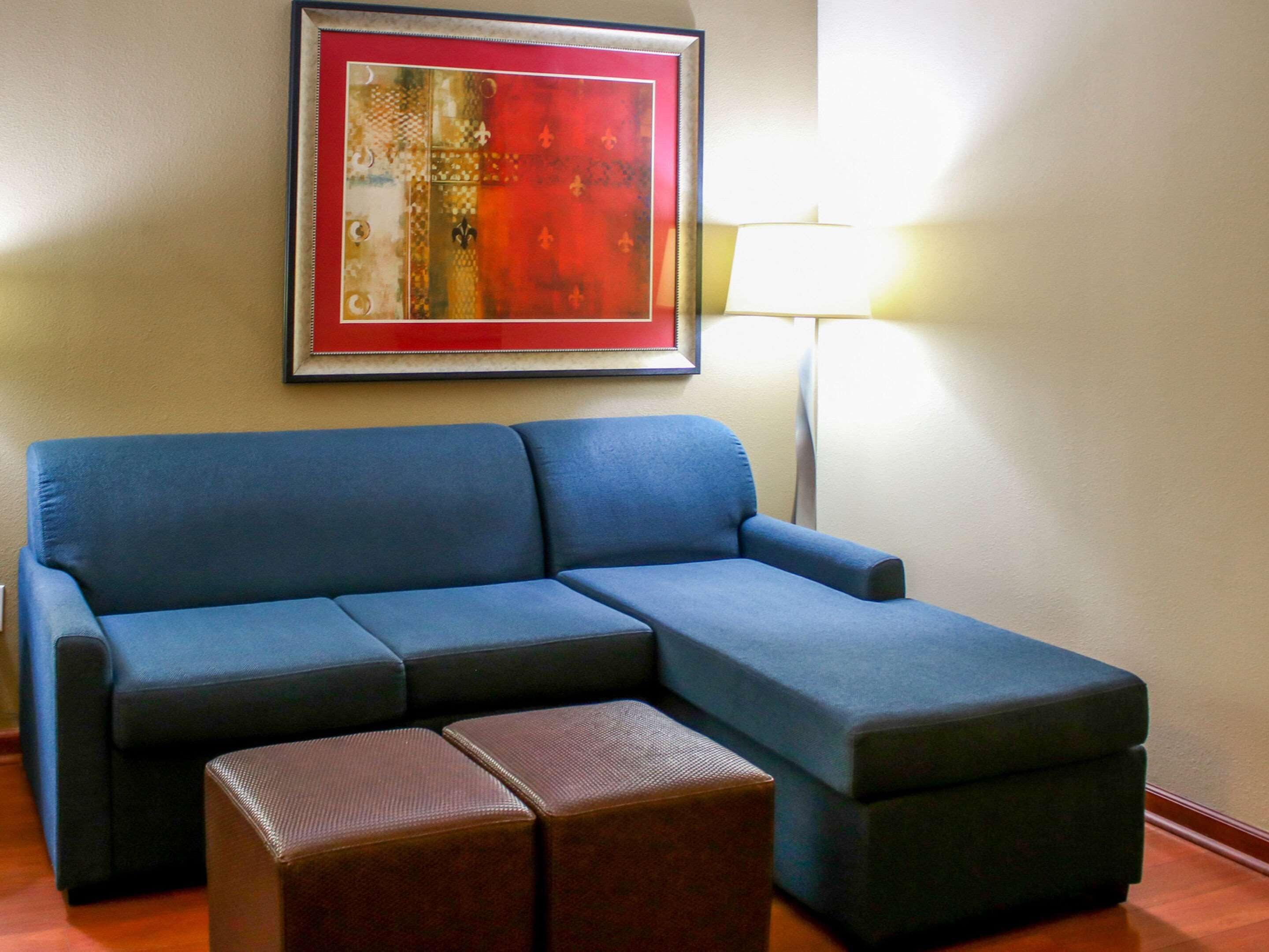Un sofá seccional azul con un reposapiés marrón al frente, una obra de arte enmarcada en la pared y una lámpara de pie a la derecha.