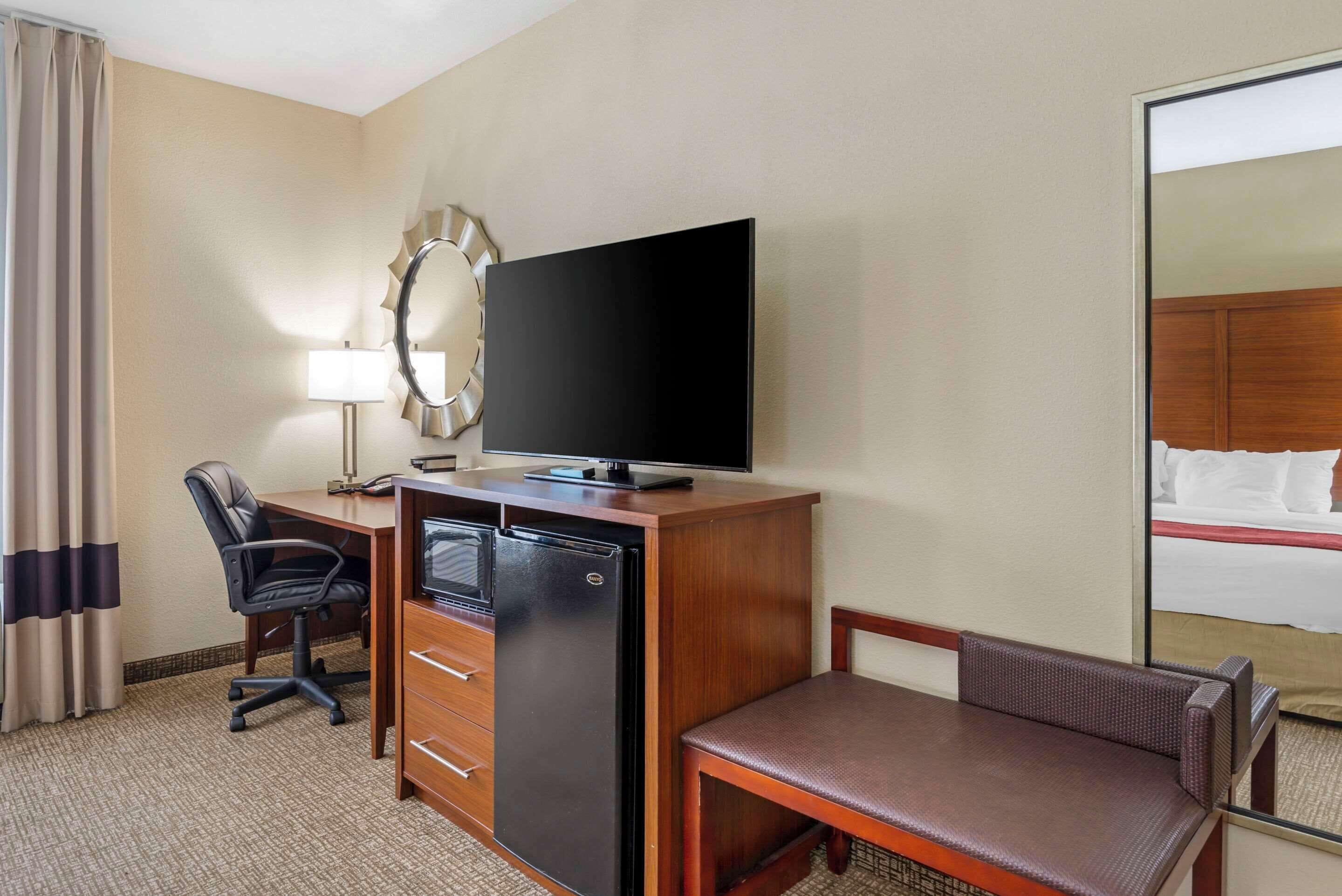 Habitación de hotel con escritorio de madera, refrigerador negro, televisor, espejo, silla y cama con ropa de cama blanca.