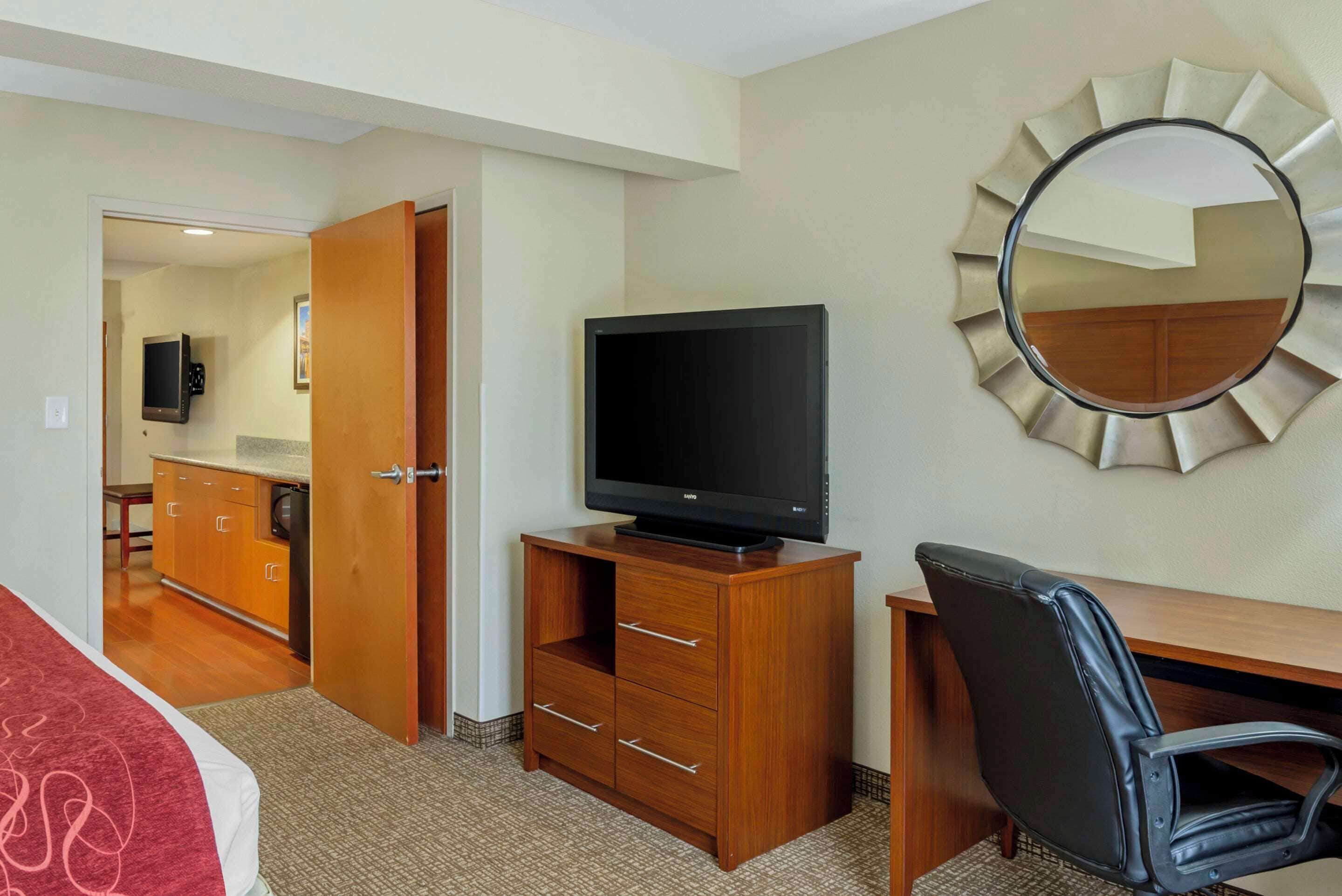 A hotel room with a wooden desk, a black leather chair, a television on a wooden cabinet, a mirror on the wall, and a door leading to a bathroom.