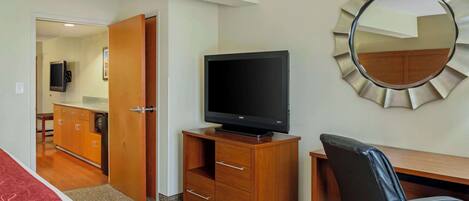 A hotel room with a wooden desk, a black leather chair, a television on a wooden cabinet, a mirror on the wall, and a door leading to a bathroom.