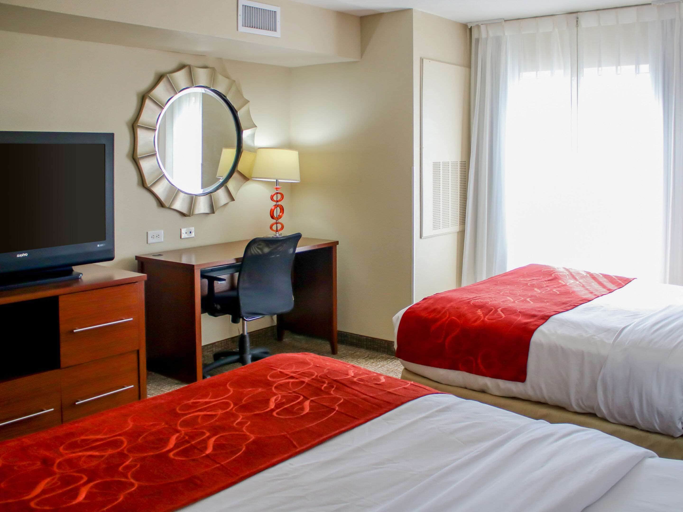 A hotel room with a TV, desk, chair, two beds with red and white bedding, a round mirror, and a window with curtains.