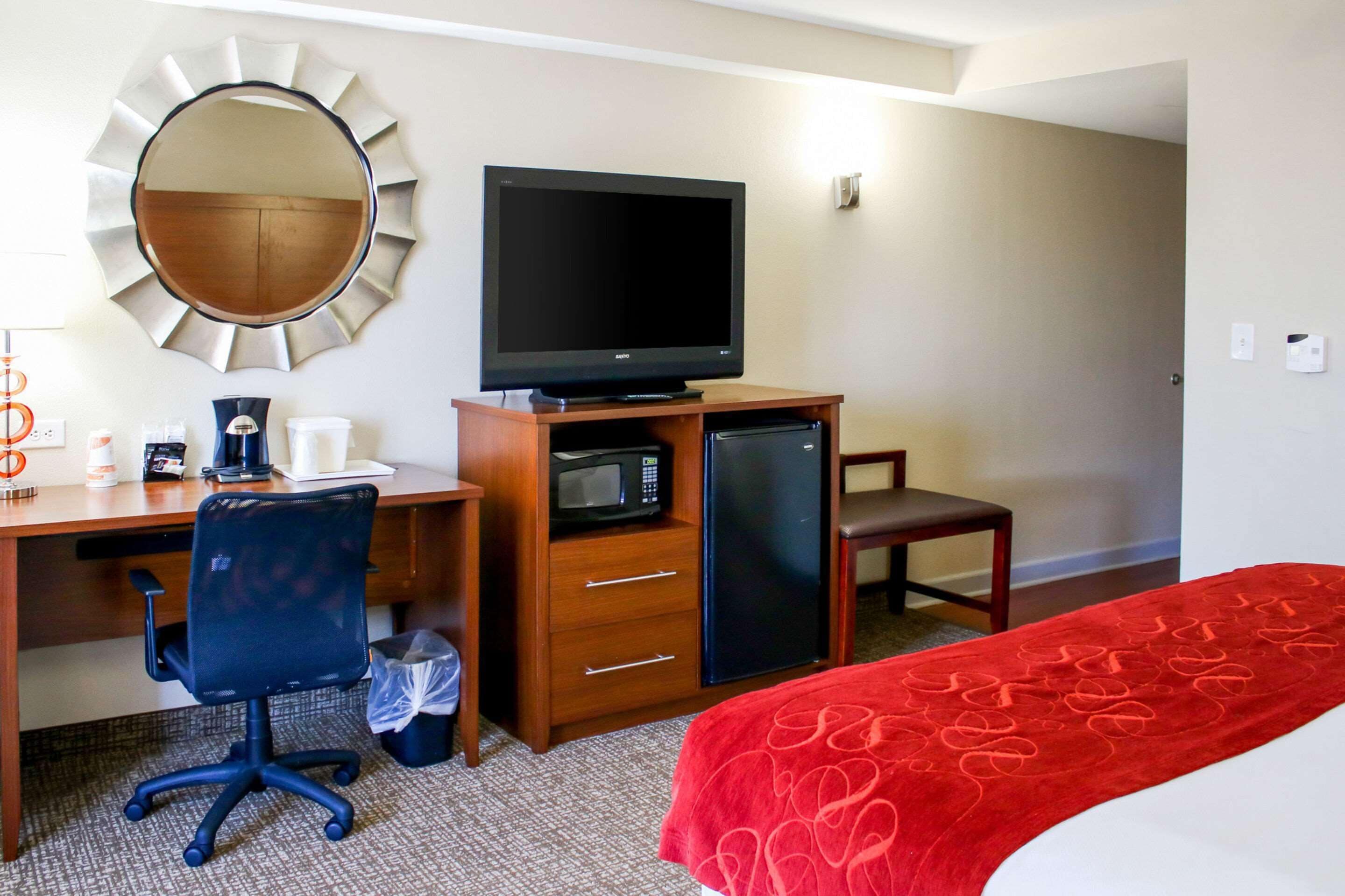 Habitación de hotel con escritorio de madera, televisor, microondas, refrigerador, una silla y una cama con manta roja.
