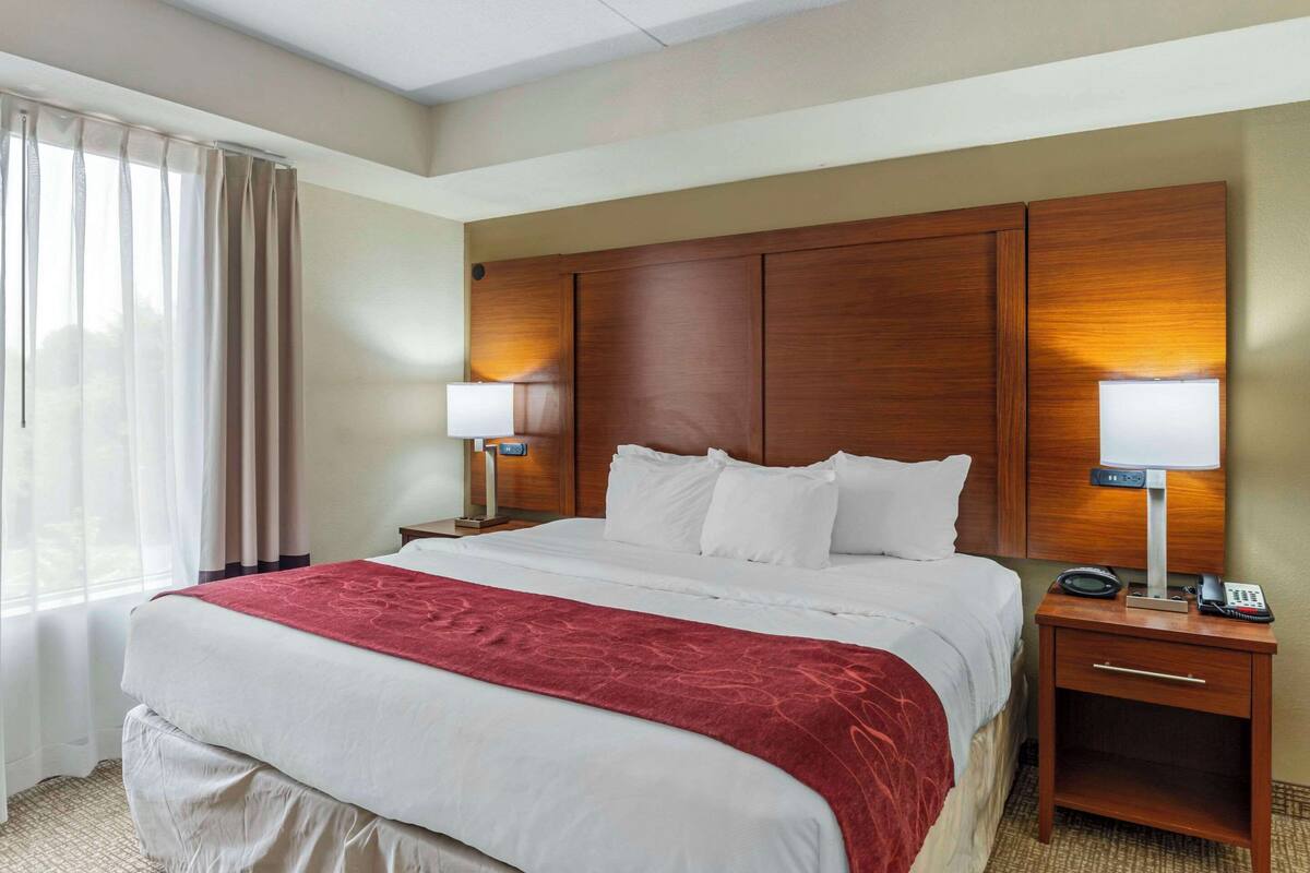 A hotel room with a large bed, two bedside tables with lamps, a wooden headboard, and a window with curtains.