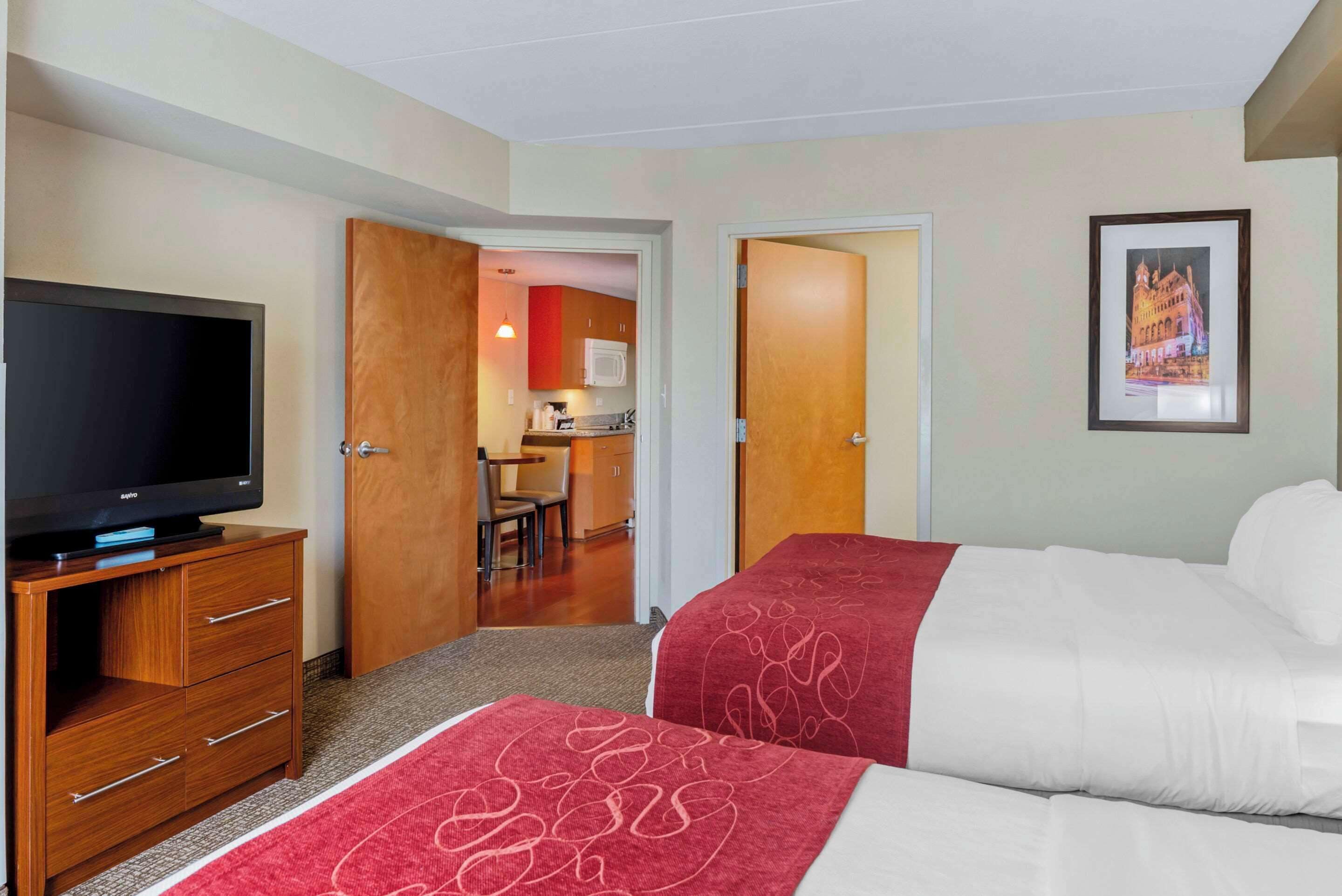 A hotel room with two beds, a TV on a wooden dresser, a kitchenette in the background, and a framed picture on the wall.