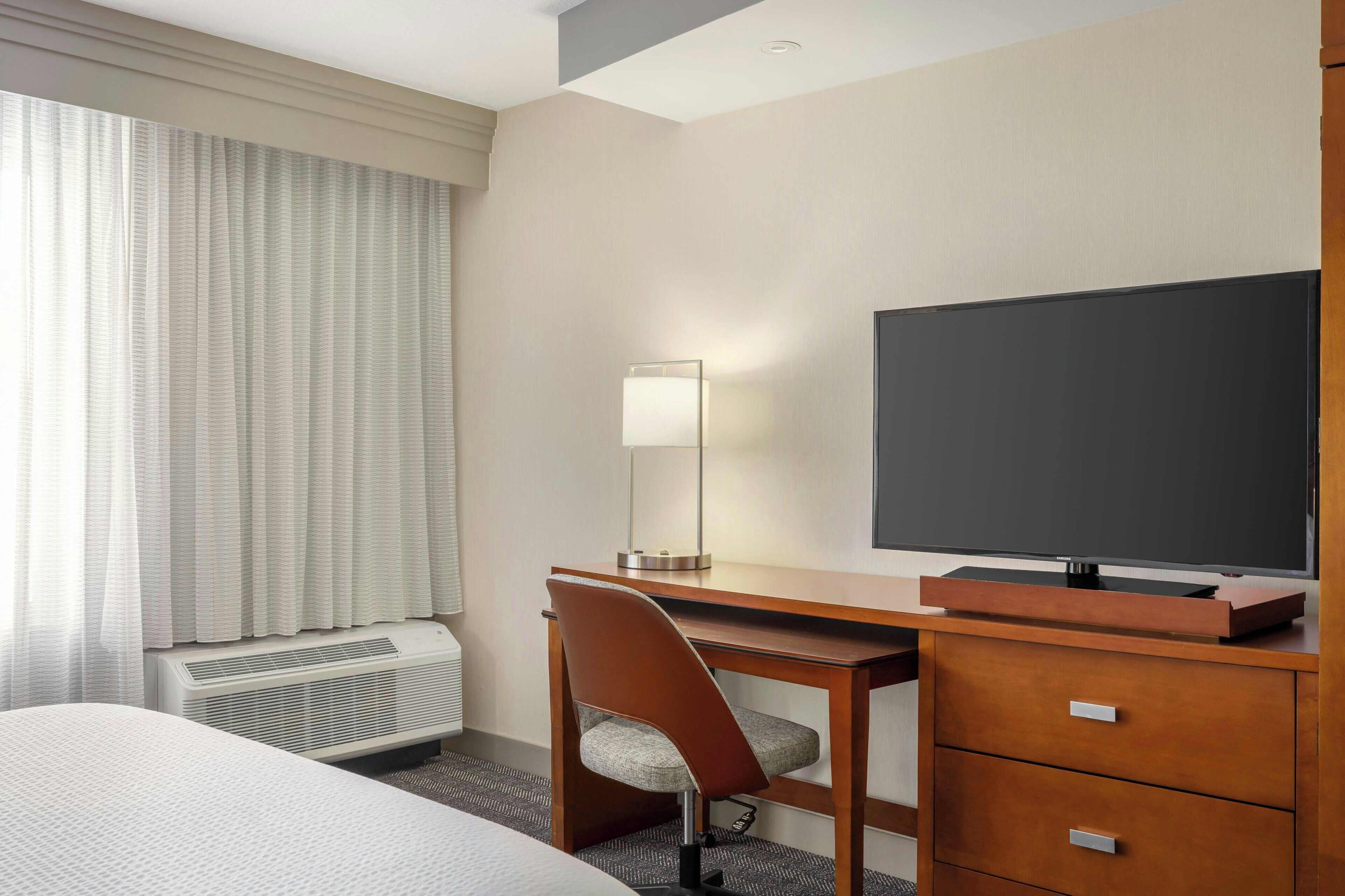 A hotel room with a bed, a desk with a chair, a television, a mirror, and a heater.