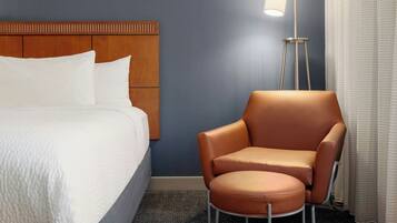 A hotel room with a bed, a brown leather chair, a floor lamp, and a window with curtains.