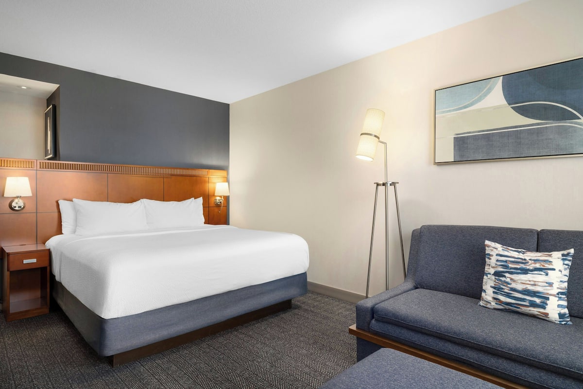 A hotel room with a large bed, a bedside table with a lamp, a sofa with a patterned pillow, and a floor lamp.