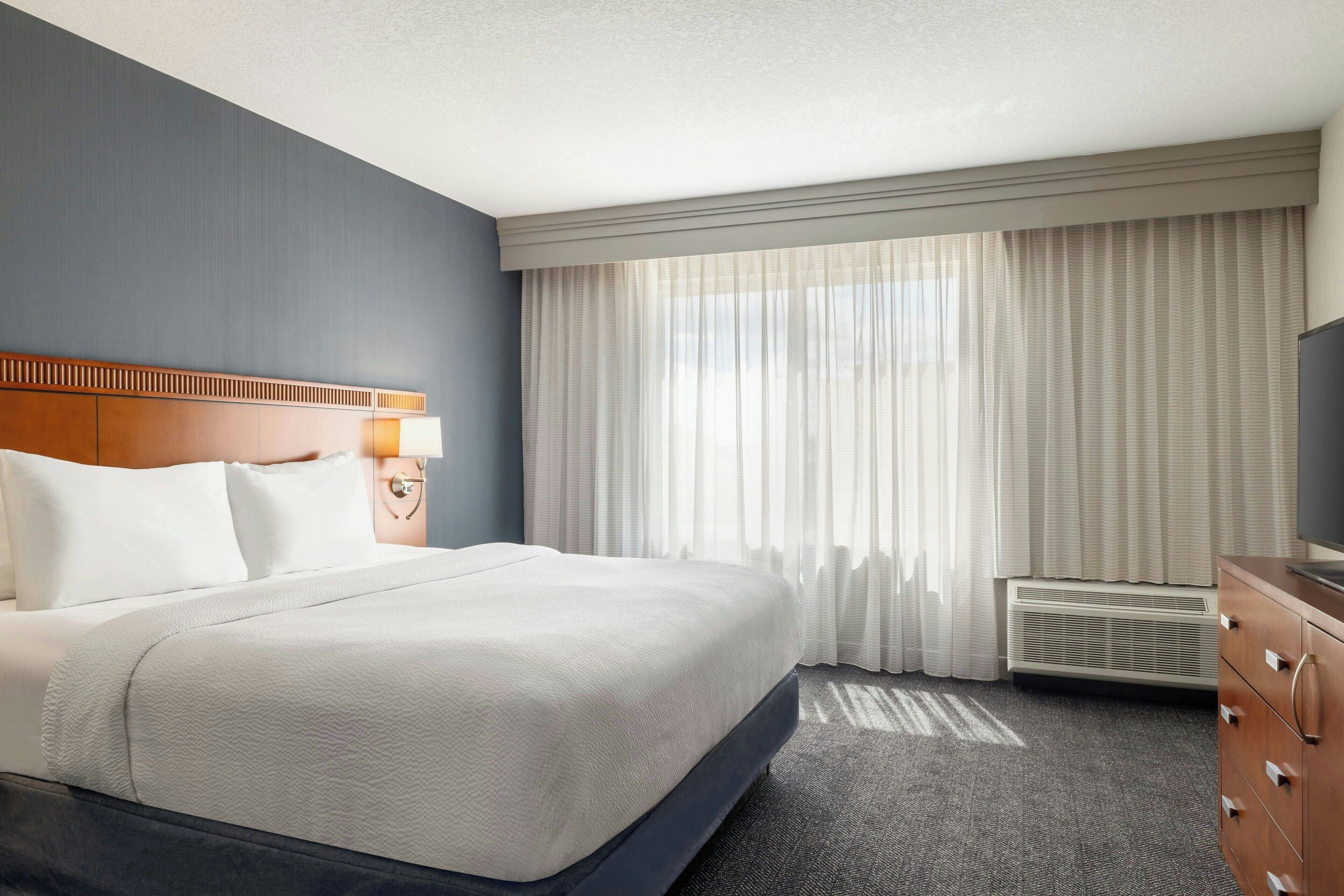 A hotel room with a large bed, a wooden headboard, a bedside table with a lamp, a dresser, and a window with sheer curtains.