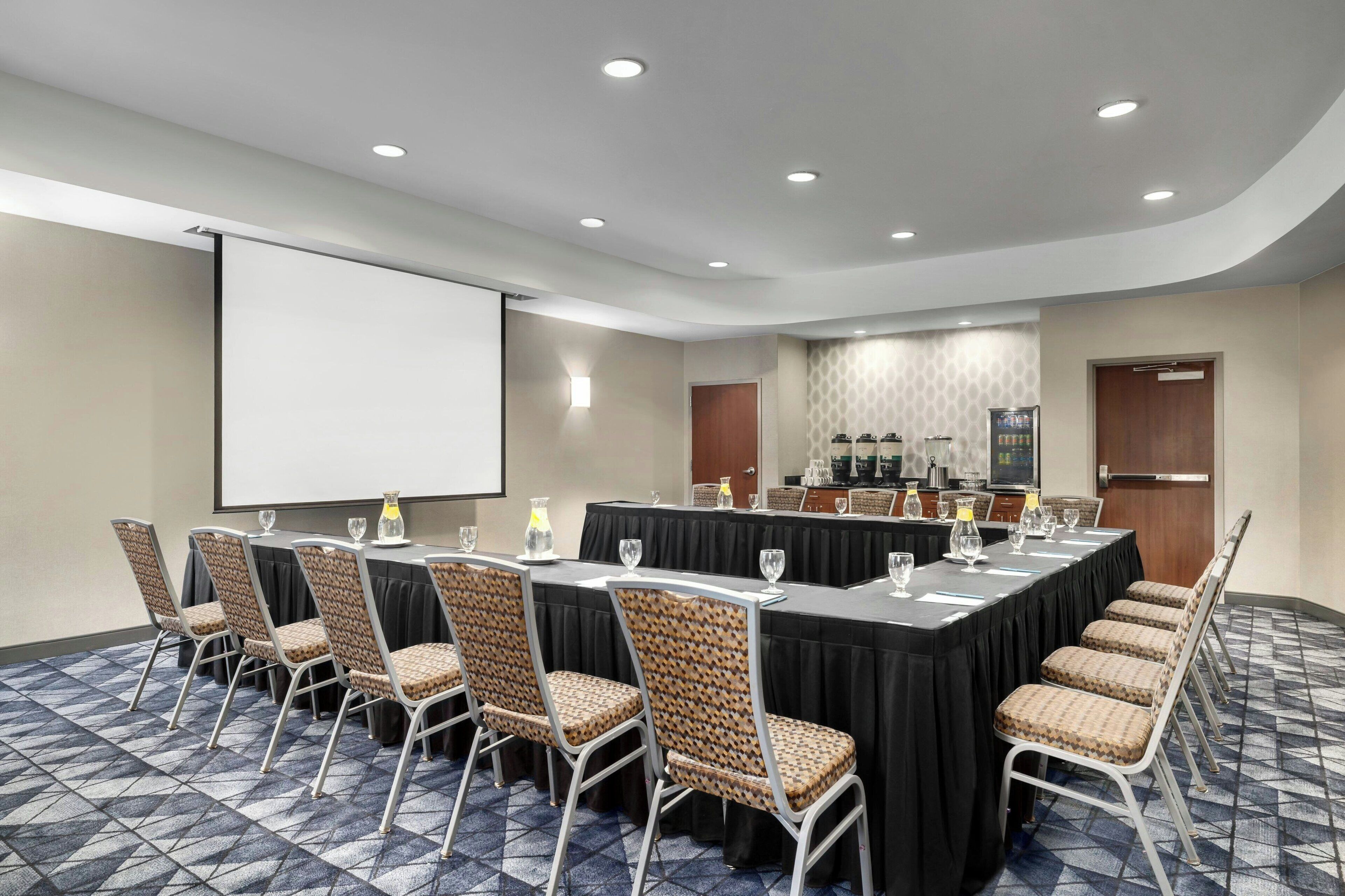 Un salón de conferencias con una larga mesa, sillas, una pantalla de proyector y una zona de buffet.