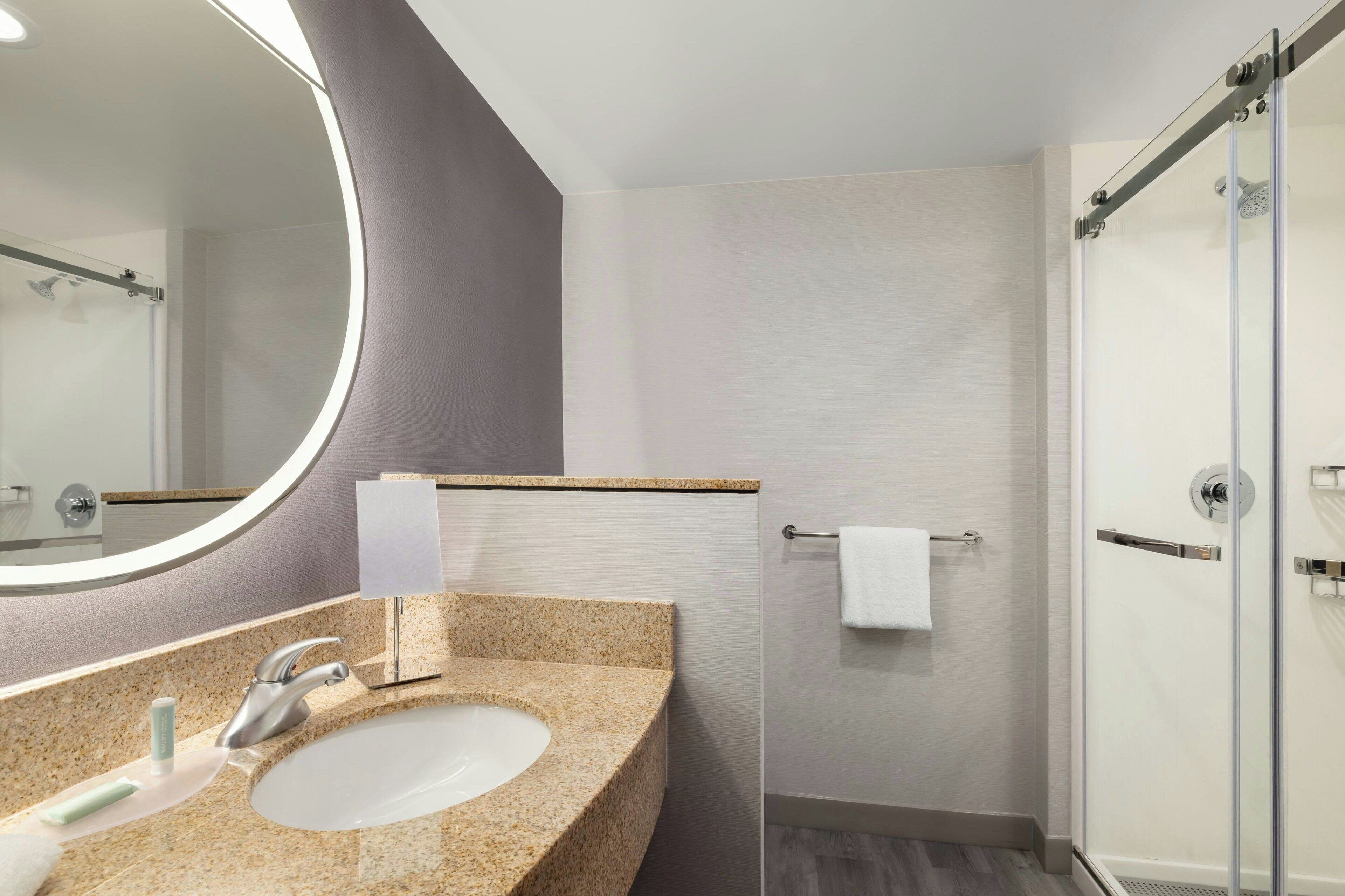 A modern bathroom with a granite countertop sink, a round mirror, a glass shower enclosure, and a towel rack.