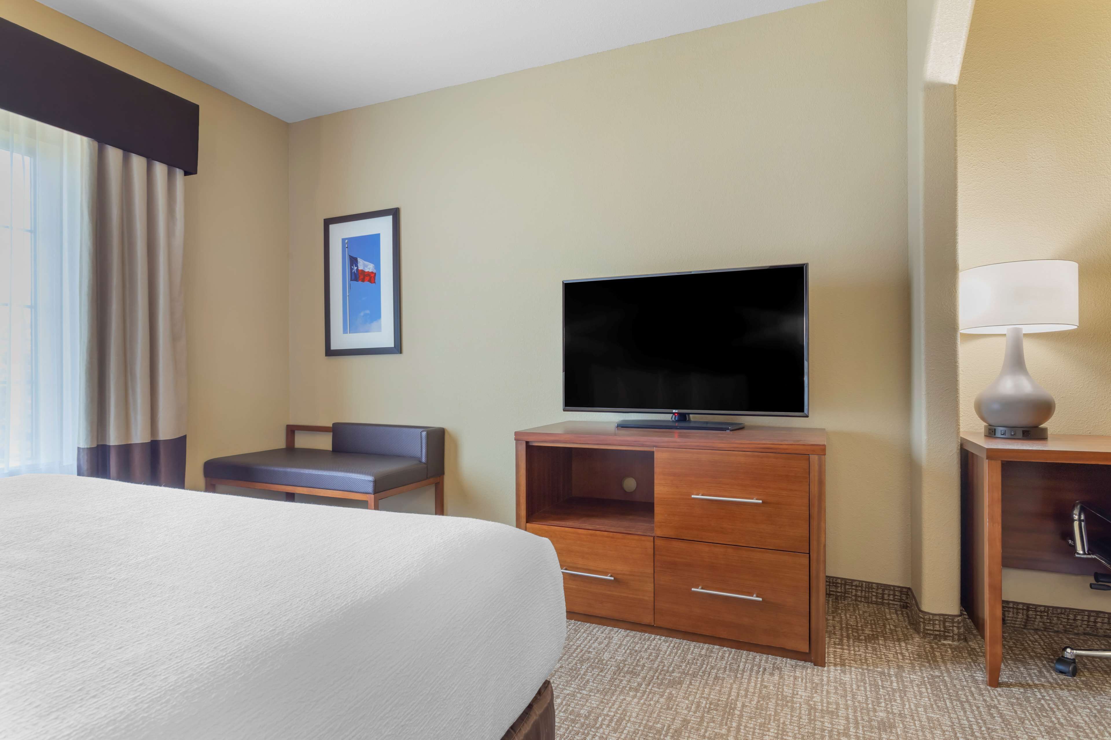A hotel room with a bed, a TV on a wooden stand, a desk with a lamp, and a framed picture on the wall.