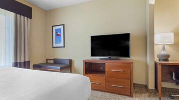 A hotel room with a bed, a TV on a wooden stand, a desk with a lamp, and a framed picture on the wall.