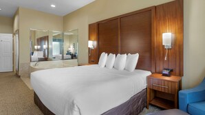 Habitación de hotel con una cama grande, cabecera de madera, mesita de noche con lámpara, espejo y una silla azul.