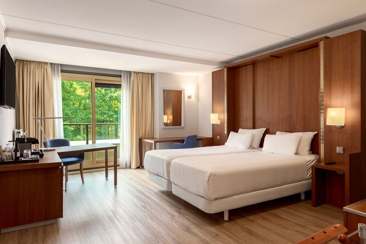 Habitación de hotel con escritorio, dos camas, televisor y un ventanal grande con cortinas.