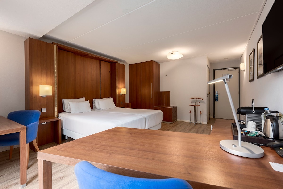 A hotel room with a bed, desk, chair, bedside table, lamp, and a television.