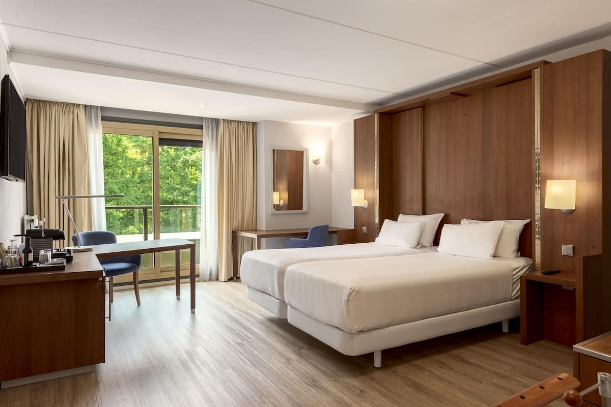 Habitación de hotel con dos camas, un escritorio, una silla y un ventanal con cortinas.