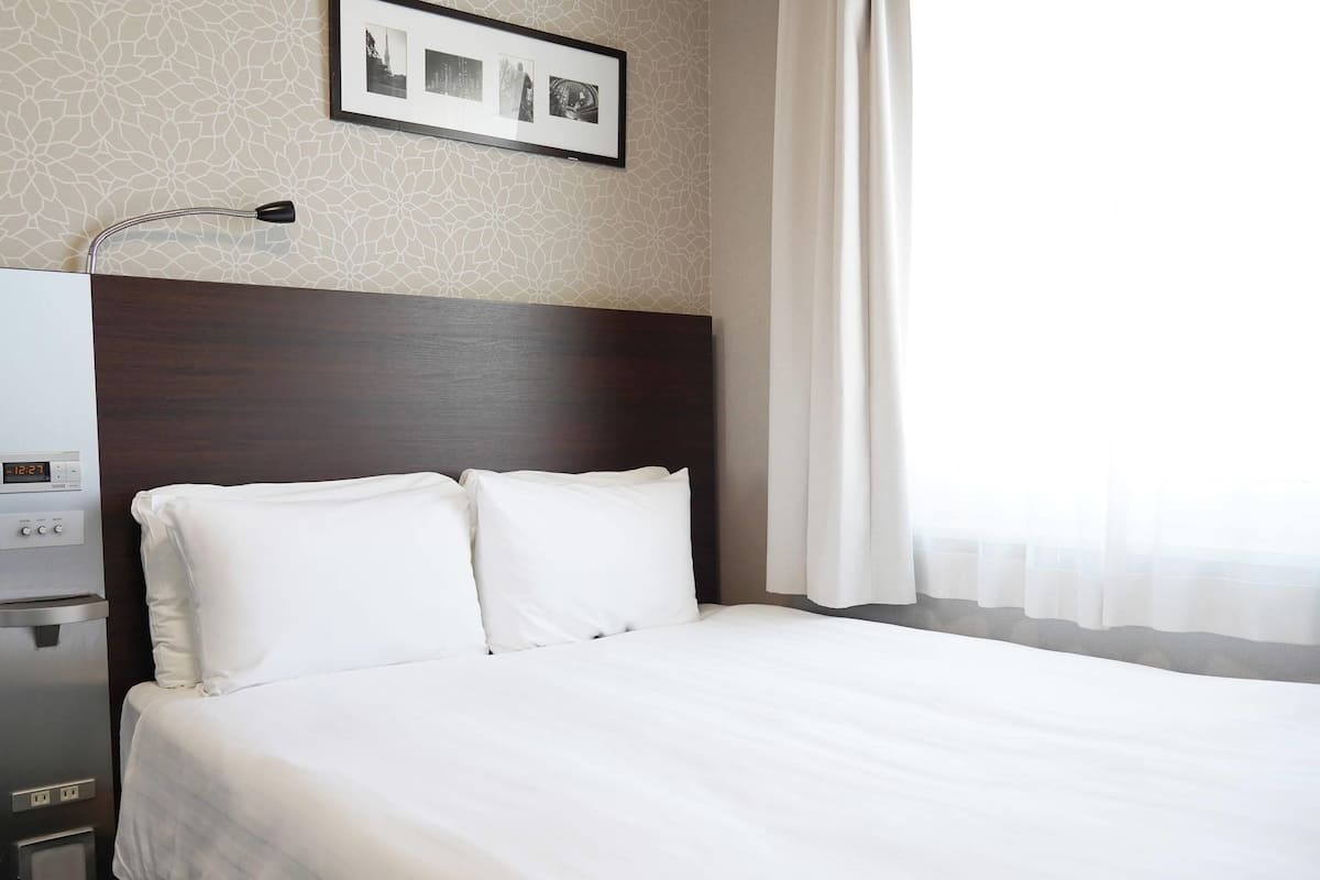 A hotel room with a large bed, a nightstand with a lamp, a framed photo on the wall, and a window with curtains.