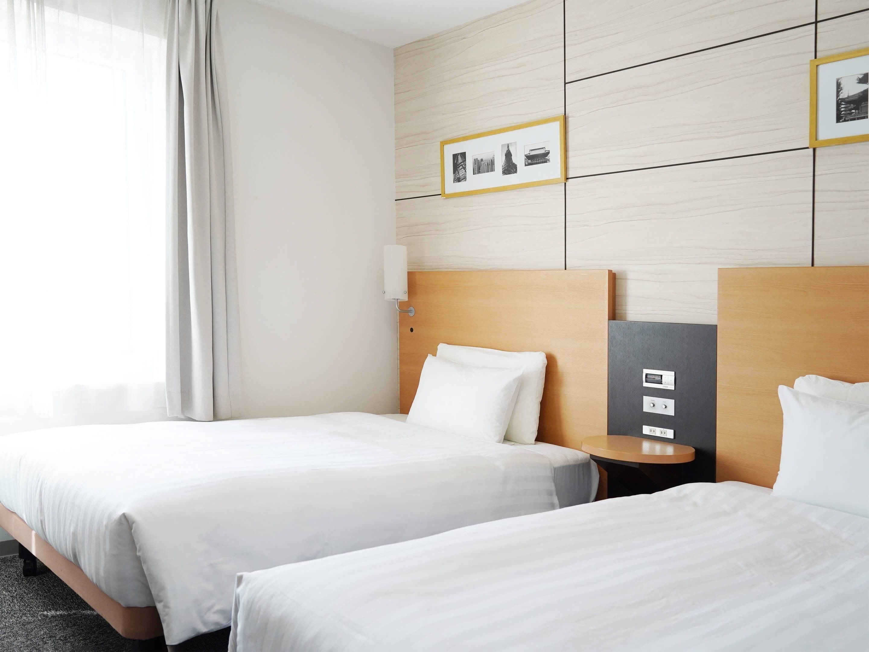A hotel room with two beds, a bedside table with a lamp, a wall-mounted light, and a framed picture above the beds.