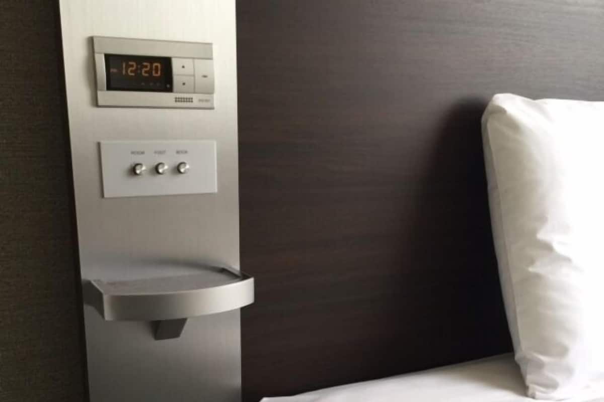 A hotel room with a bed, a digital clock, and a control panel with buttons.
