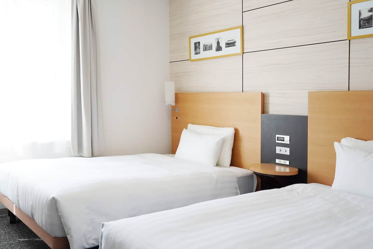 A hotel room with two beds, a bedside table with a lamp, a wall-mounted light, and a framed picture above the beds.