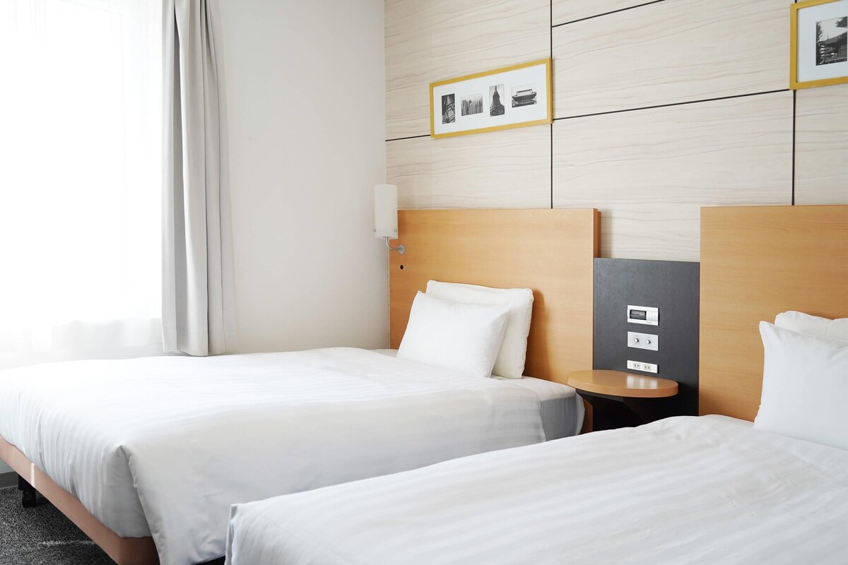 A hotel room with two beds, a bedside table, a wall-mounted light, and a framed picture above the bed.