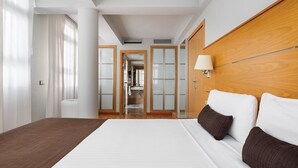 Ein Hotelzimmer mit einem großen Bett, einem hölzernen Kopfteil, einer Nachttischlampe und Blick ins Badezimmer durch eine Glastür.
