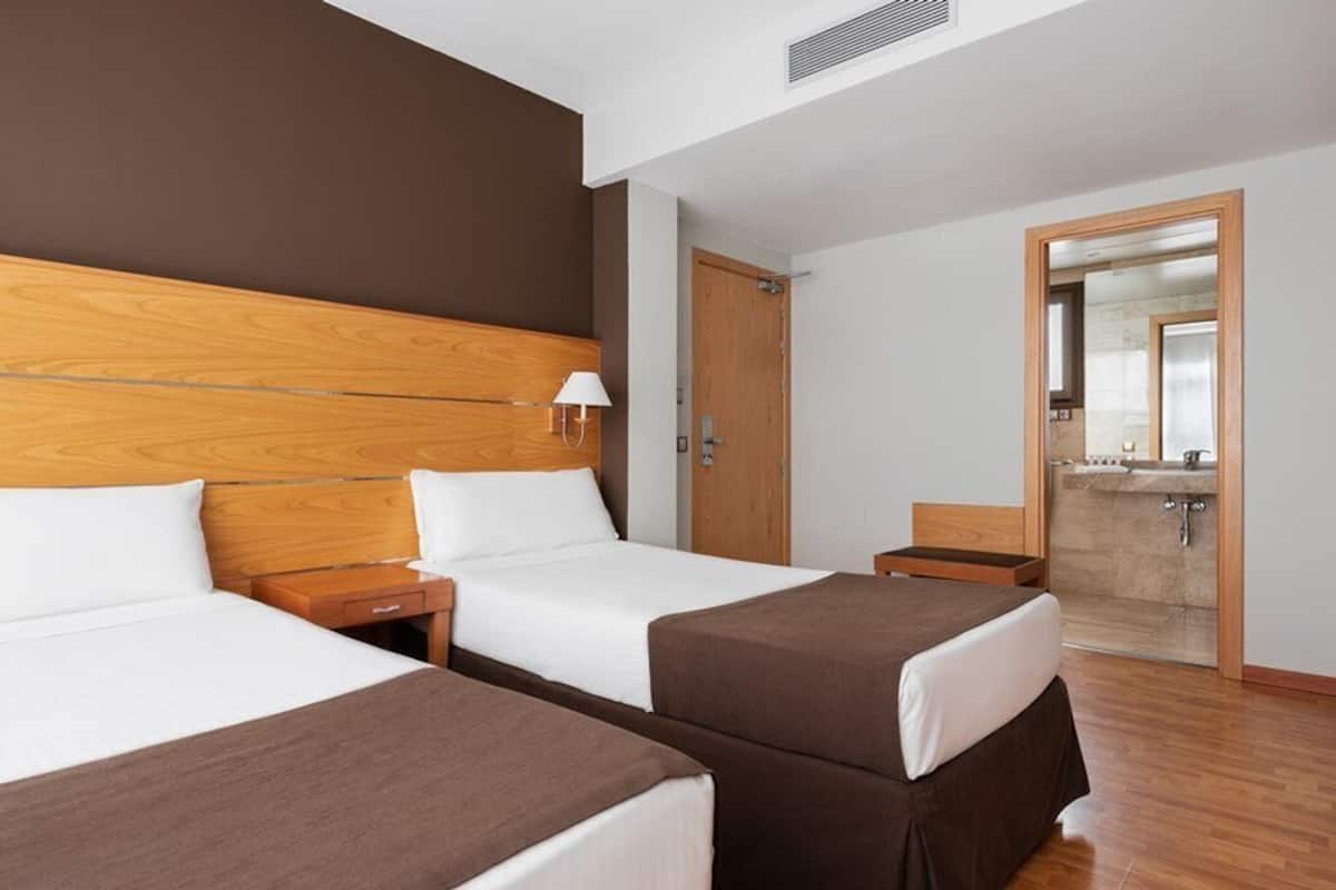 Ein Hotelzimmer mit zwei Betten, einem Holzkopfteil, einem Nachttisch mit Lampe, einem Holzboden und einem durchgehenden Türspalt sichtbaren Badezimmer.