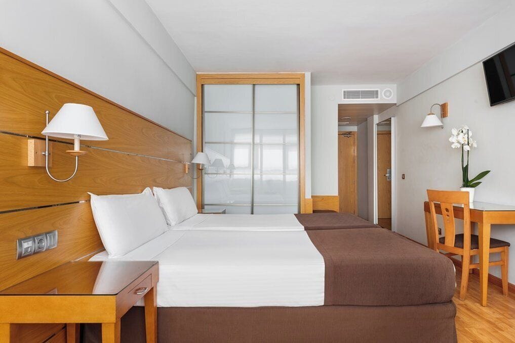 Una camera d'albergo con una testiera di legno, un letto con biancheria da letto bianco e marrone, una scrivania con una sedia, una TV a parete e un'orchidea in vaso sulla scrivania.