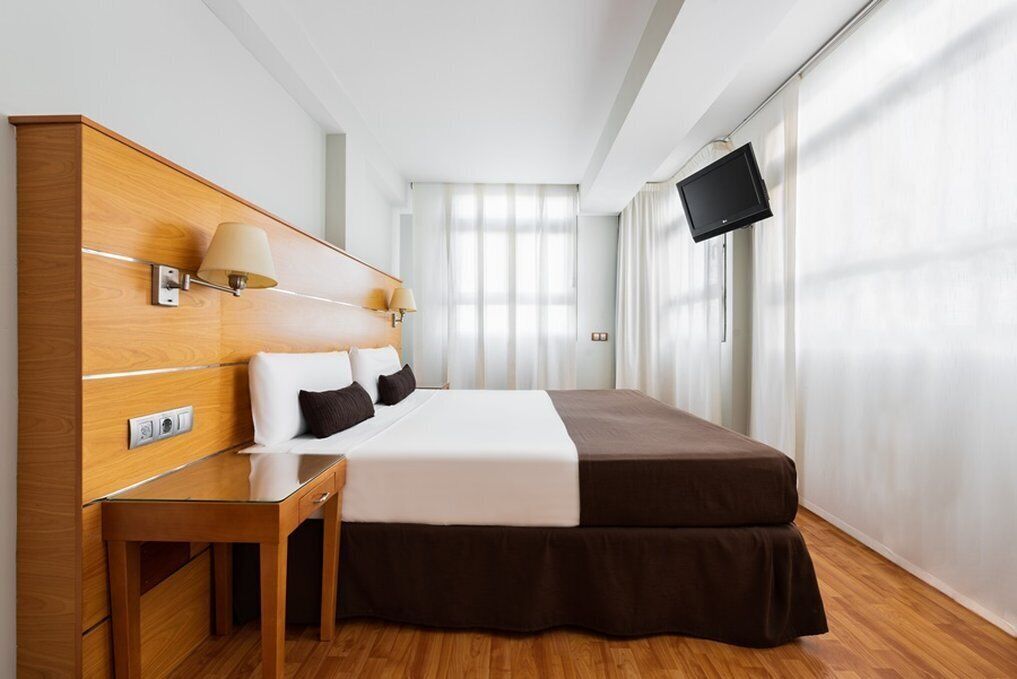 A hotel room with a wooden headboard, a bed with white and brown bedding, a bedside table with lamps, a flat-screen TV mounted on the wall, and a large window with sheer curtains.