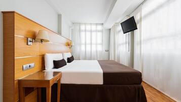 Habitación de hotel con cabecera de madera, cama con ropa de cama blanca y marrón, mesita de noche con lámparas, televisor de pantalla plana en la pared y un gran ventanal con cortinas traslúcidas.