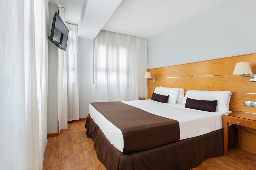 Une chambre d’hôtel avec un grand lit, une tête de lit en bois, un téléviseur fixé au mur et une table de chevet avec une lampe.
